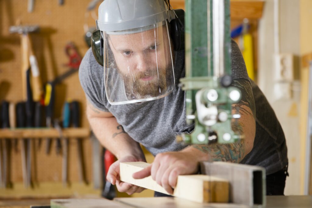 Craftsman with safety mask visor handles band saw in workshop